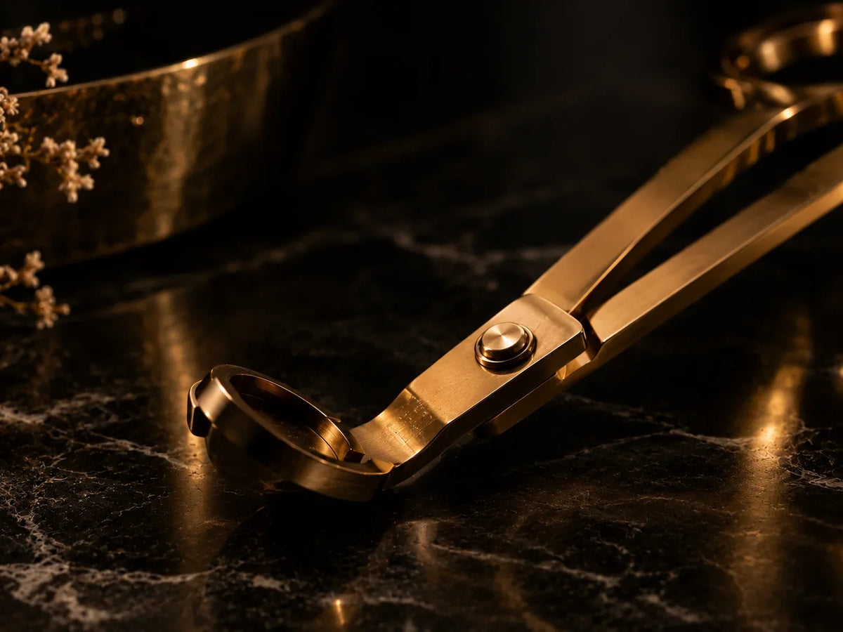 Goldener Kerzendochtschneider in Nahaufnahme auf schwarzem Marmor mit feinen Maserungen und warmen goldenen Lichtreflexionen. Die dunkle, elegante Inszenierung unterstreicht die hochwertige Ästhetik des Kerzenzubehörs.
