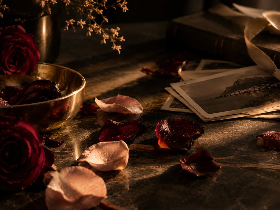 Dunkelrote und rosafarbene Rosenblätter auf einer dunklen Marmoroberfläche neben alten Fotografien und einer goldenen Schale. Warmes, goldenes Licht erzeugt eine sinnliche, nostalgische Atmosphäre als Sinnbild für Duft und Erinnerung.